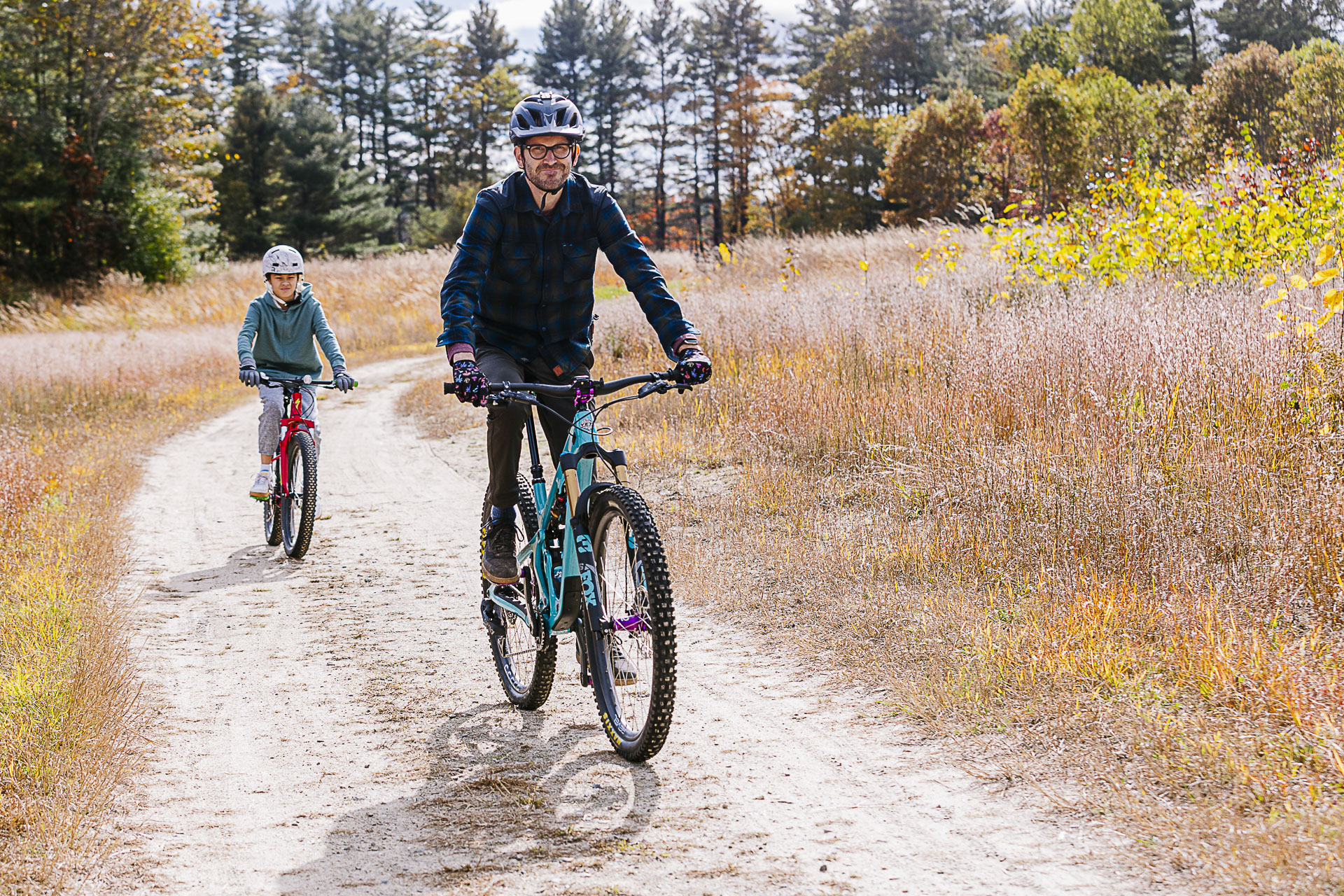 An adult and a young rider pedal side by side down a sandy fire road through autumn grasses