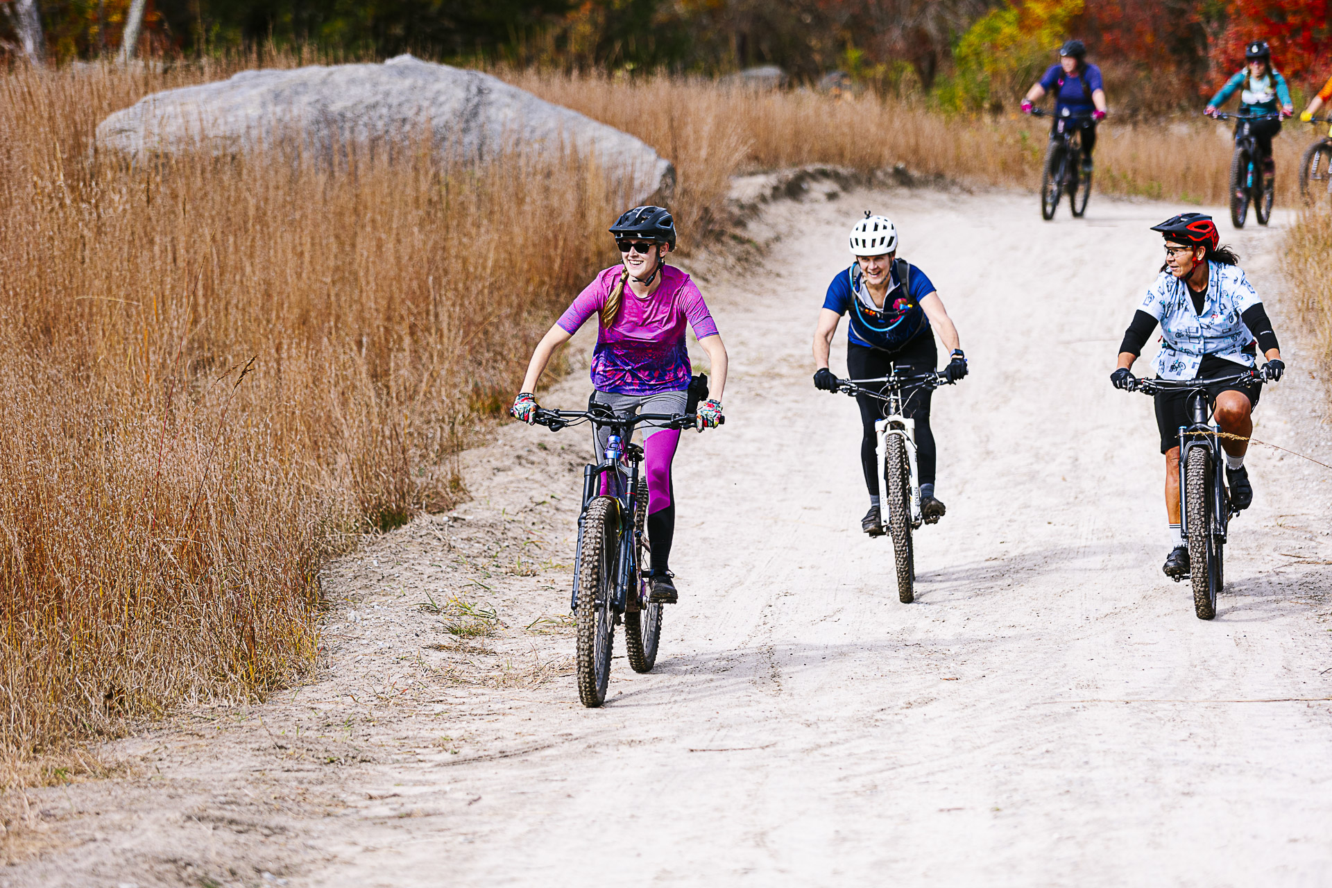 Three riders smile as they roll toward the camera on a wide sandy trail, with more riders behind them