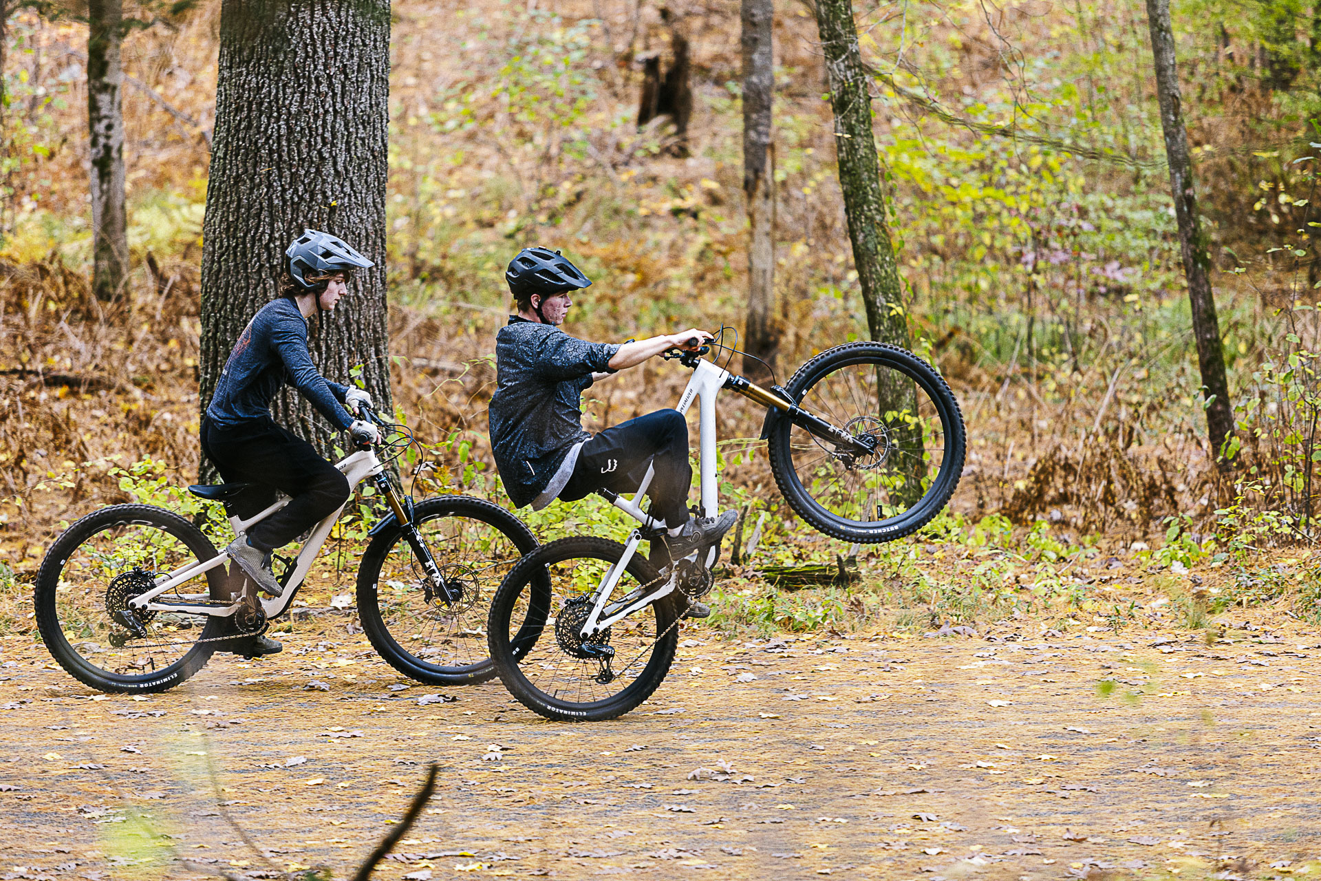 Two young riders on a wooded trail, one lifting his front wheel into a wheelie