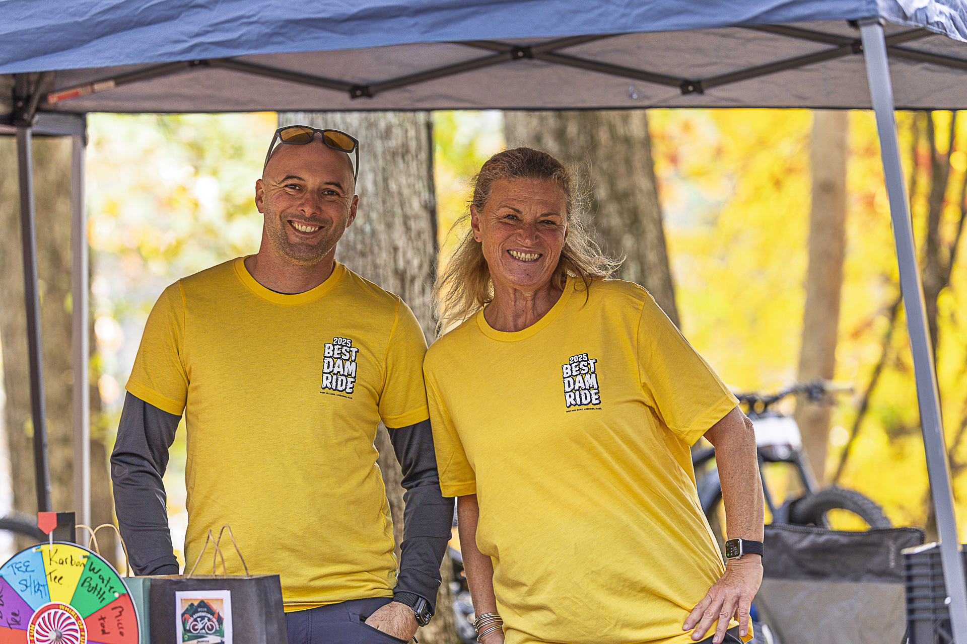 Two volunteers in yellow Best Dam Ride shirts smile behind the raffle table and prize wheel