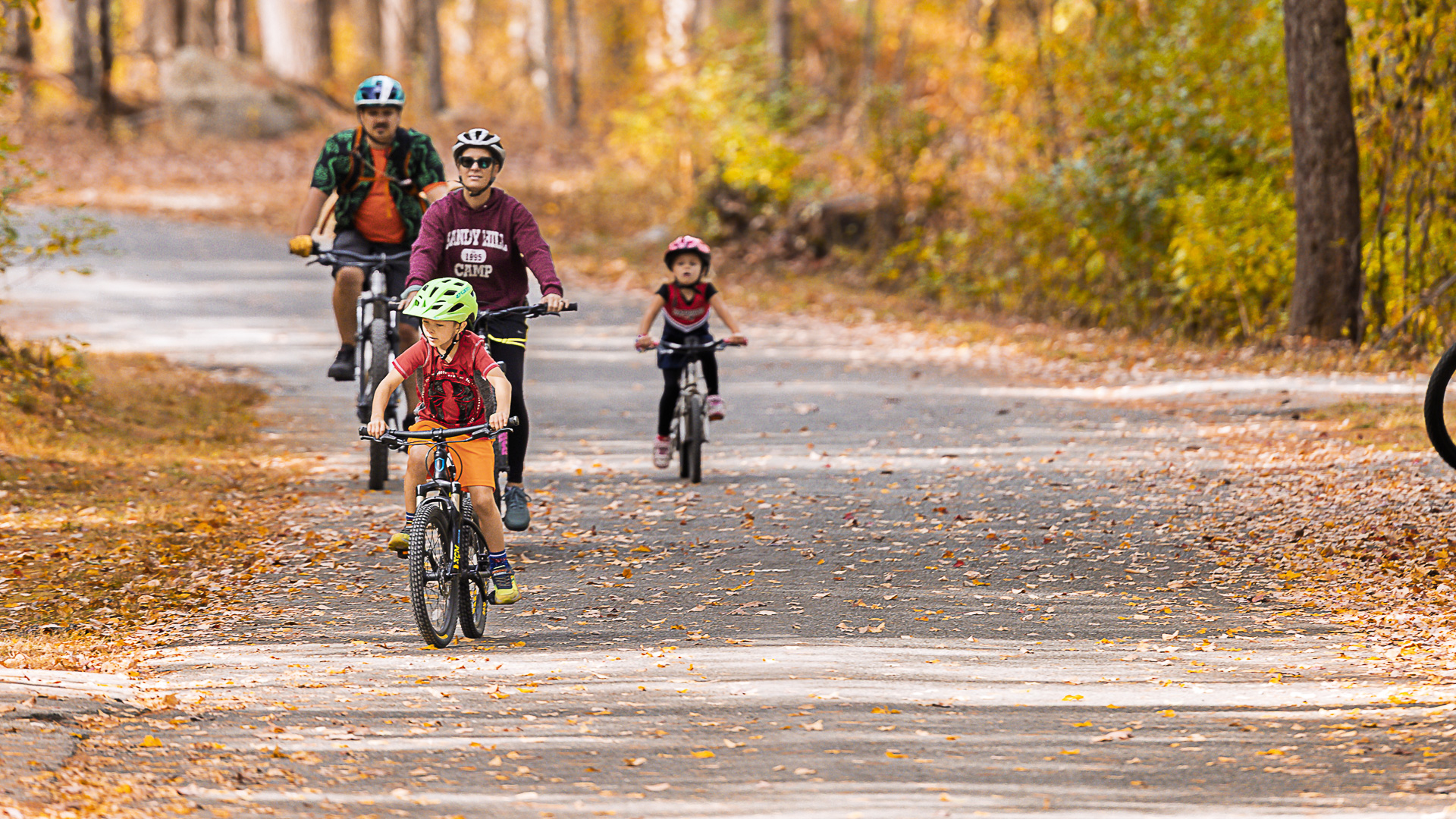 A family rides together down a leaf-covered road, two young kids out front