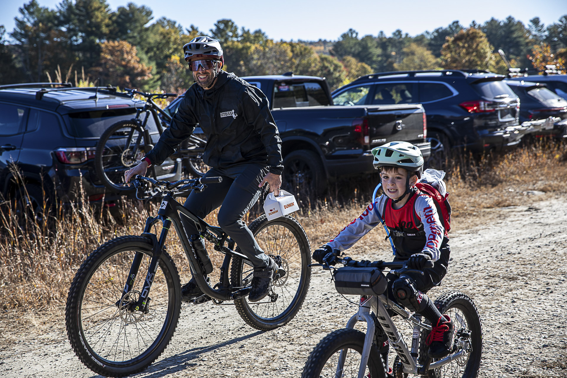 An adult and a young rider grin as they pedal out along a gravel road
